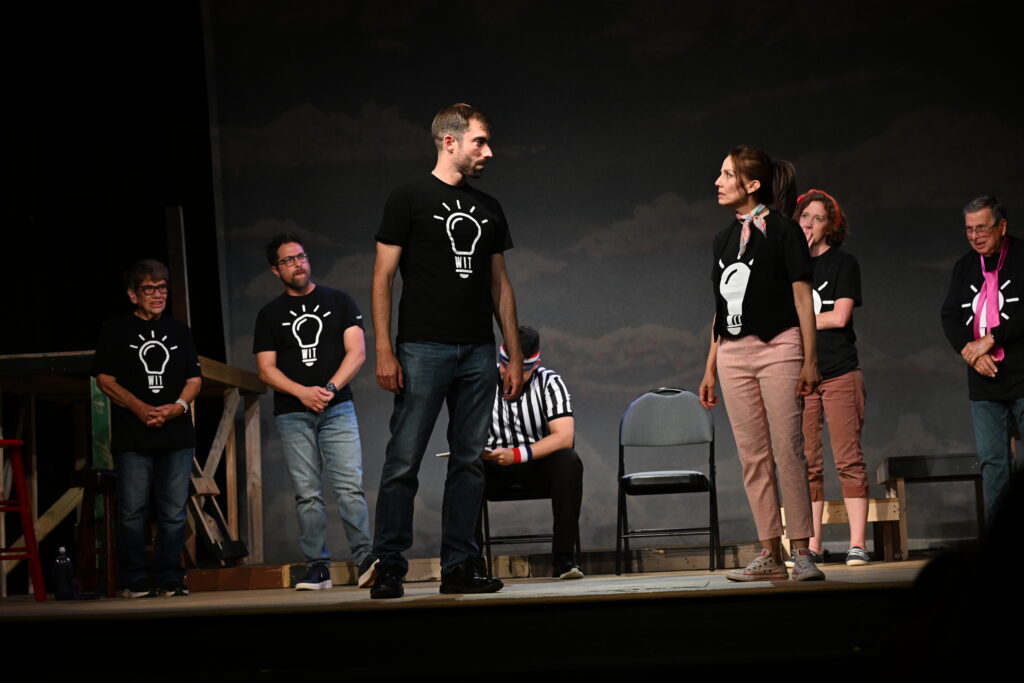 Two WIT! performers face to face in a scene while the rest of the troupe watches